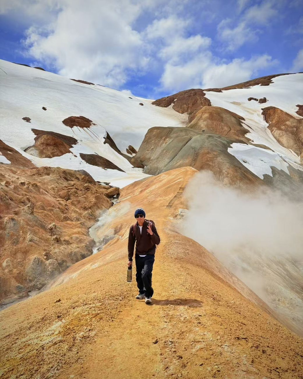 Ethan Chen walking a ridge of ochre coloured ground between snowfields, with steam rising beside the trail.